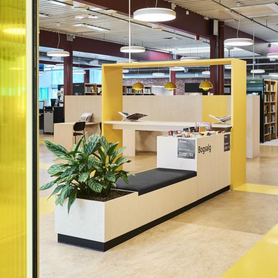 Fotografi taget ind gennem bibliotekets gule skydedøre. I midten ses en plantekasse, en læsebar med udstillede bøger, samt bogreoler.