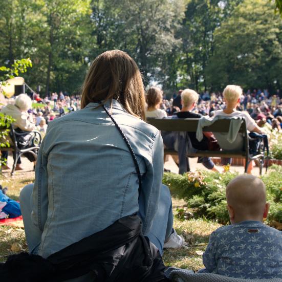 En kvinde, der sidder på en græsplæne med ryggen til kameraet. Ved siden af hende ses et lille barn, og foran dem en større menneskemængde, der også sidder på græsset og lytter til musik.