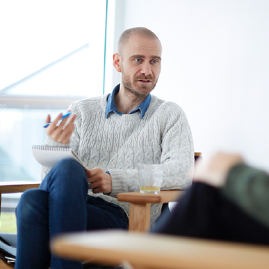 Mand, der sidder i en stol. Han gestikulerer, og snakker med en kvinde, der sidder med siden til i forgrunden.