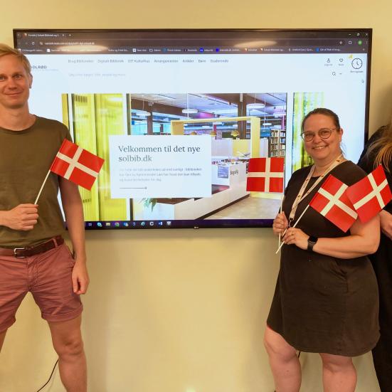 Bibliotekets systemadministratorgruppe (Rasmus, Lisbeth og Fie) foran en skærm med den nye hjemmeside på. De holder alle tre små flag og smiler.