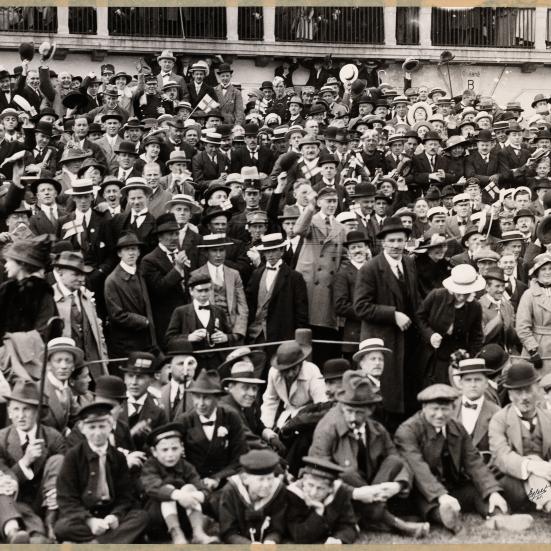 Fotografi i sort / hvid af en stor gruppe mennesker. De er alle nydeligt klædt på, og bærer hatte. De er til fodboldkamp udenfor, og står i tilskuerrækker.