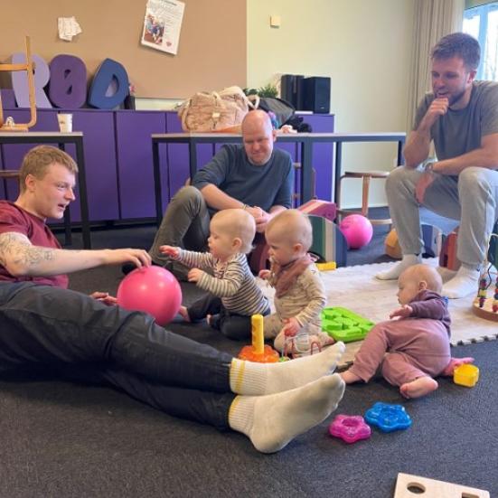 En gruppe små børn, der leget på gulvet mede en masse farverigt legetøj. Ved siden af dem sidder tre fædre og leger med.