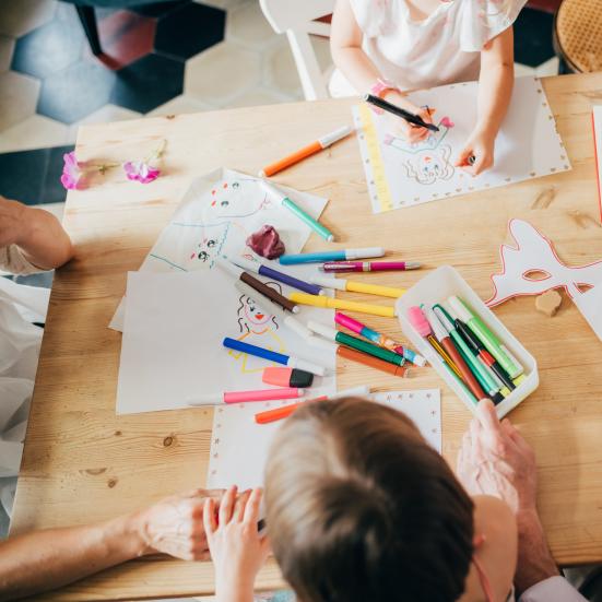 Et bord med børn rundt om. På bordet er papir og farvestrålende tuscher. Børnene tegner, mens en voksen kigger på.