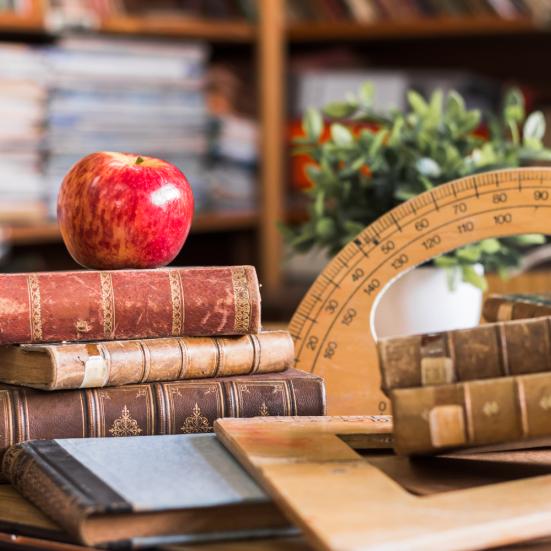En stak bøger, der ligger foran en lineal på et bord. Der står et æble på stakken af bøger, og i baggrunden ses en plante og fyldte reoler.