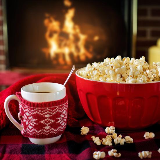 Krus med varm kakao med et rødt omslag på, ved siden af en rød skål fyldt med popcorn. Det står på et rødt tæppe foran en tændt pejs.