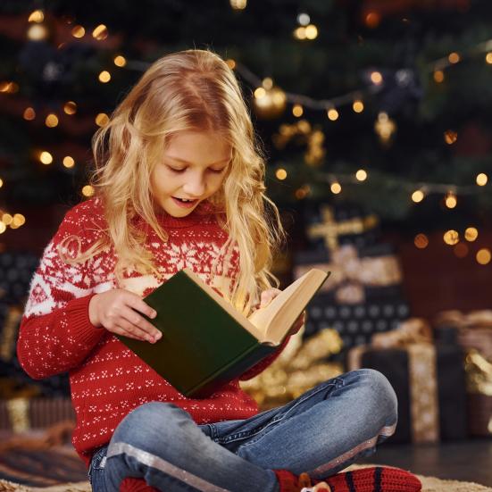 Pige iført julesweater, der læser en bog foran et juletræ.