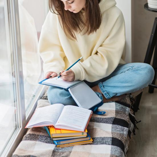 Kvinde sidder i en vindueskarm og skriver noter i en notesbog. Foran hende ligger en stak bøger.