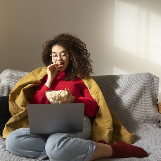 Kvinde, der sidder i en sofa med tæppe på. Hun ser film på sin laptop og spiser popcorn.