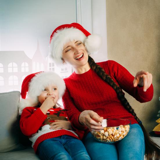 En kvinde og et barn sidder i en sofa foran et juletræ og ser film. De er begge iført nissehue.