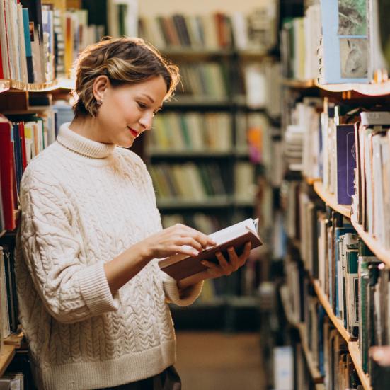 Kvinde, der kigger i en bog i et bibliotek. Hun står mellem reoler fyldt med bøger.