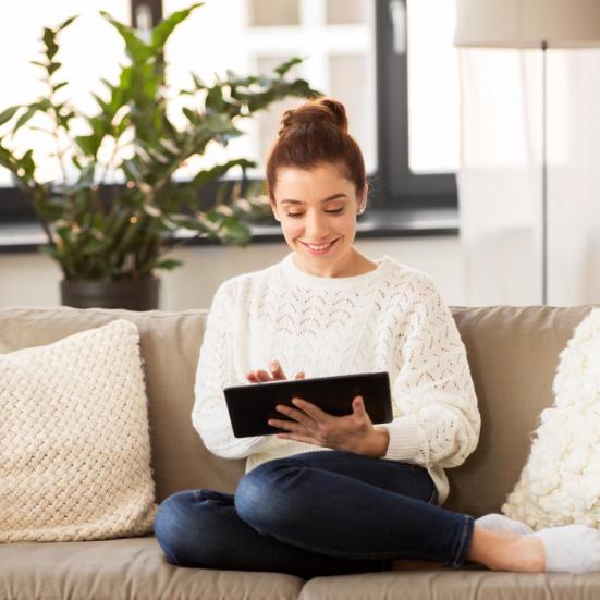 Kvinde sidder i en sofa med en tablet. Hun smiler, og sidder i en hyggeligt stue.