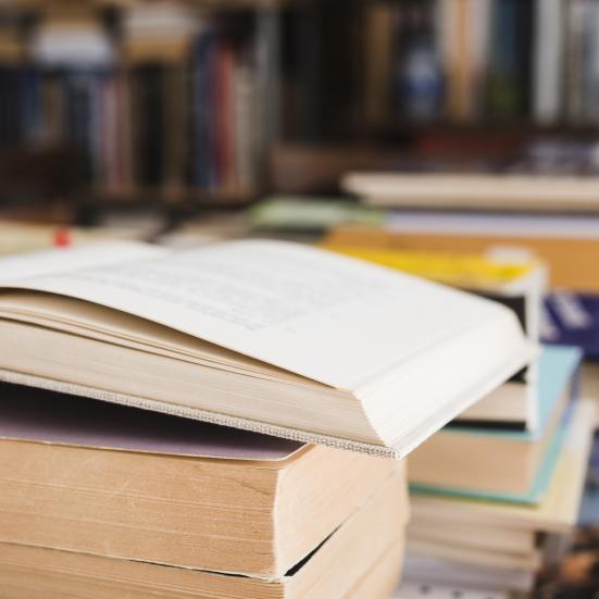 En åben bog, der ligger på en stabel af bøger. I baggrunden ses fyldte bogreoler.