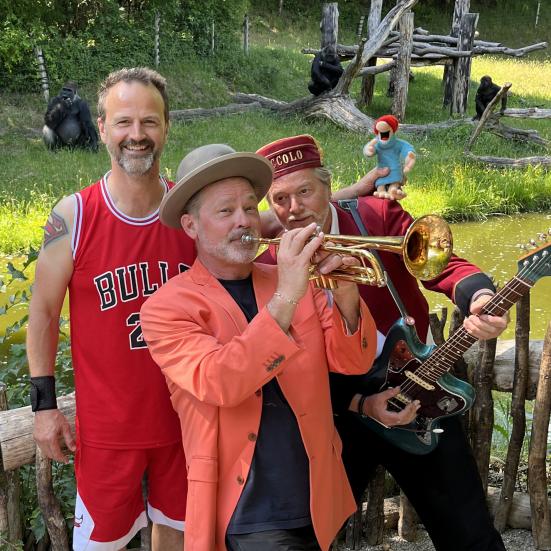 Et orkester bestående af tre mænd i rødligt tøj. Den forreste mand spiller på trompet og har en hat på. 