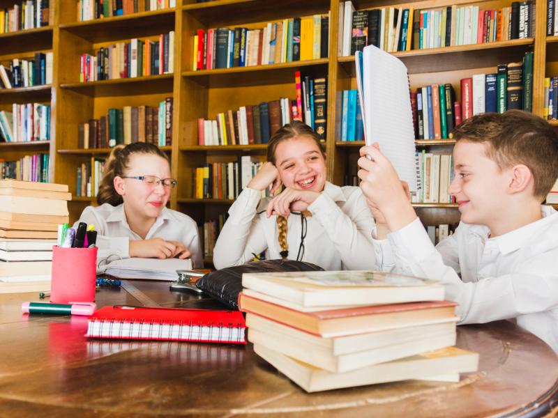 Tre børn sidder og snakker sammen ved et bord i et bibliotek. Den ene holder en notesbog, mens de andre kigger og griner. På bordet ses bøger, notesbøger og skriveredskaber.