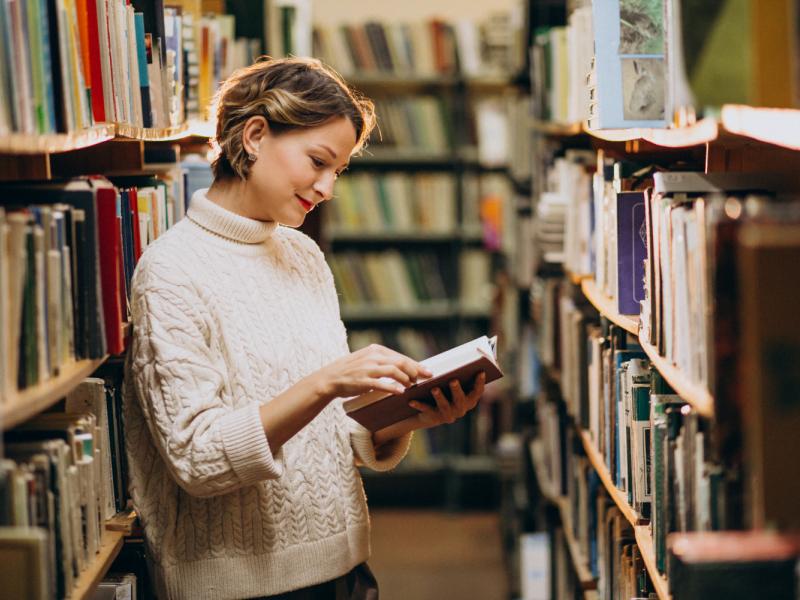 Kvinde, der kigger i en bog i et bibliotek. Hun står mellem reoler fyldt med bøger.