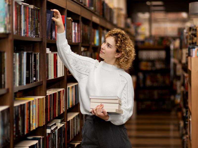 Kvinde, der er ved at tage en bog fra en reol i et bibliotek. Hun holder en stor stak bøger.