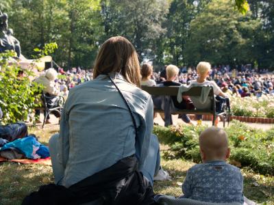 En kvinde, der sidder på en græsplæne med ryggen til kameraet. Ved siden af hende ses et lille barn, og foran dem en større menneskemængde, der også sidder på græsset og lytter til musik.