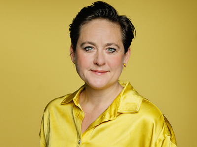 Portrætfoto af forfatter Sara Alfort på sennepsgul baggrund. Hun har en gul skjortebluse på, og kigger ind i kameraet med et lille smil.