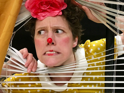 Kvinde med klovnesminke, der skærer ansigter og kigger gennem persienner. Hun er iført en gul, polkaprikket bluse med hvid flæsekant, og en stor, lyserød blomst på hovedet.