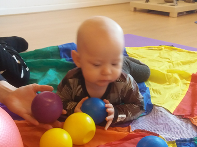 En baby på et regnbuefarvet legetæppe, der leger med små bolde. Til venstre sidder en voksen og leger med.