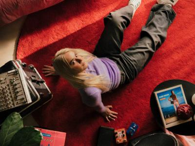 Sanger Ella Augusta set oppefra. Hun sidder på et rødt gulvtæppe omgivet af ting.