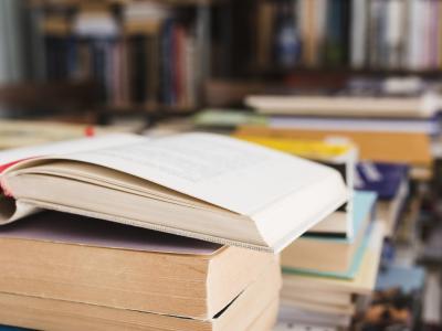 En åben bog, der ligger på en stabel af bøger. I baggrunden ses fyldte bogreoler.