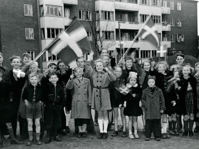 Foto i sort/hvid af en masse børn, der smiler og vifter med dannebrogsflag foran en lejlighedsbygning.