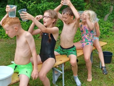 Fem børn sidder på en bænk i badetøj. De leger med vand i vandglas. 