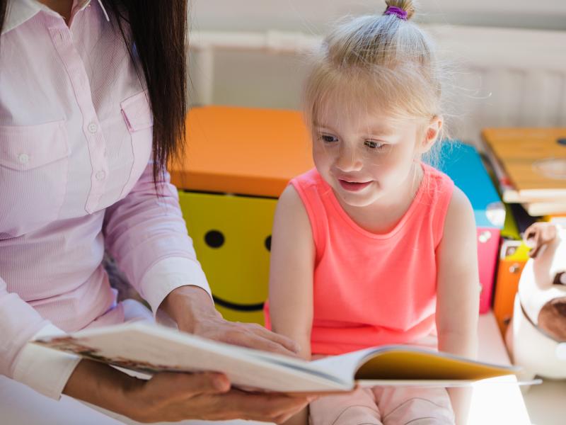 Kvinde sidder på gulvet og læser højt for et barn. Hun peger ned i en bog, mens barnet kigger med. Der er legetøj i baggrunden.