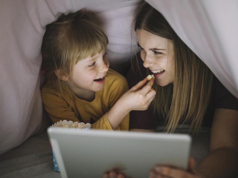 En ung pige og et barn ligger under en dyne og ser film sammen. Barnet giver pigen popkorn, mens de griner.