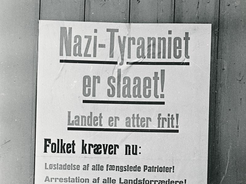 Foto i sort/hvid med plakat. Overskriften lyder "Nazi-Tyranniet er slaaet, landet er atter frit".