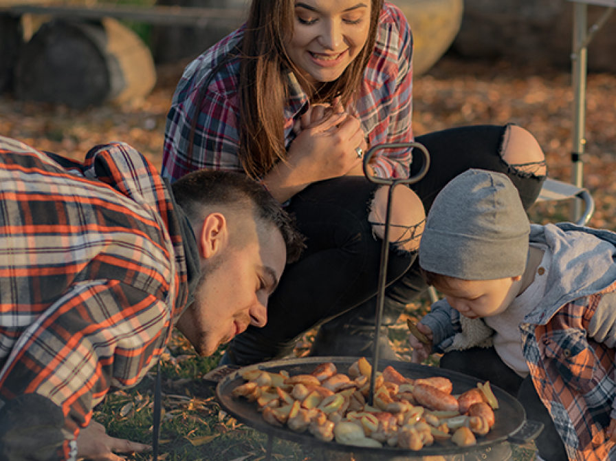 En kvinde iført en ternet skjorte, sidder på hug bag et bålfad. En mand nærstuderer maden sammen med et mindre barn.
