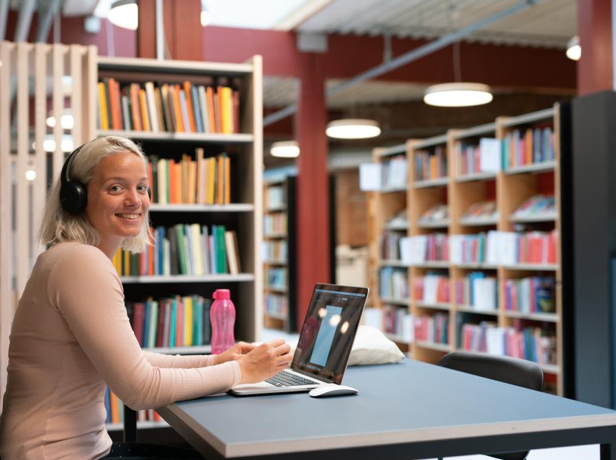 Foto Lasse Peter Nielsen. Ung kvinde studerer i biblioteket. Hun sidder ved et bord med sin computer og en lyserød vandflaske, og smiler til kameraet. I baggrunden ses hylder med bøger.