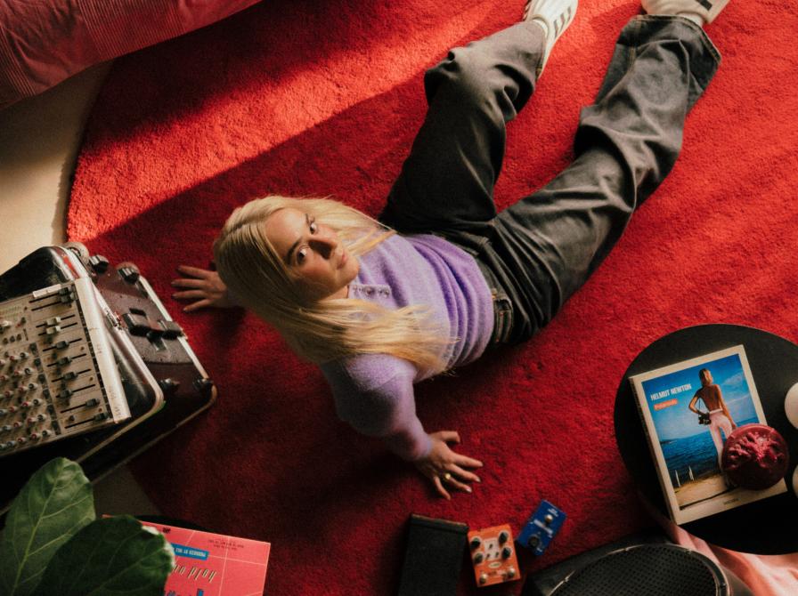 Sanger Ella Augusta set oppefra. Hun sidder på et rødt gulvtæppe omgivet af ting.