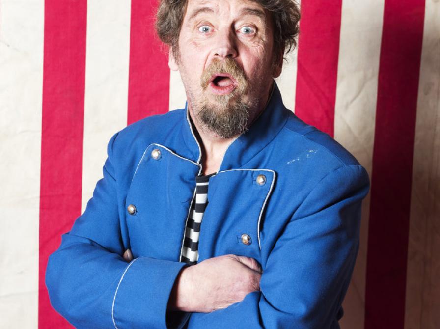 En mand i kort artist-agtig jakke sidder med åben mund foran en baggrund med røde og hvide striber. 